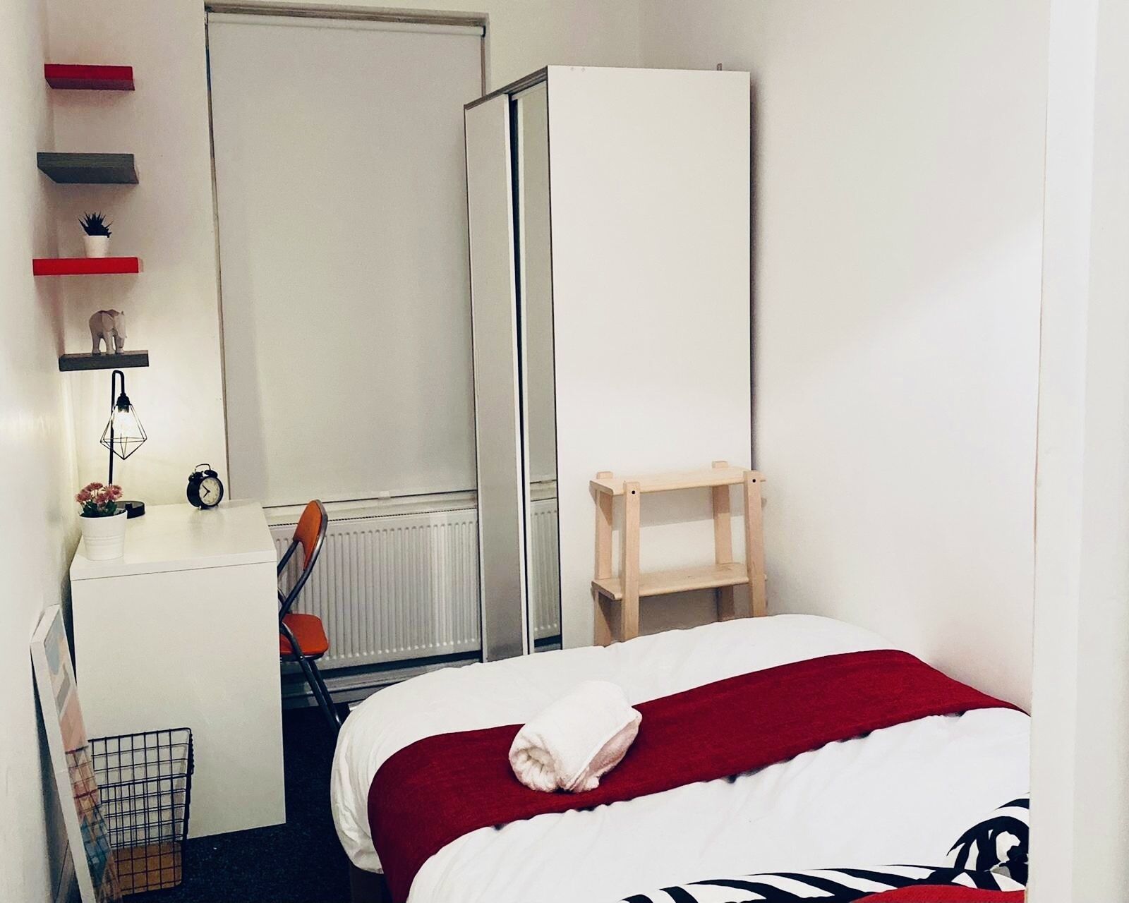 A bedroom with a bed, a radiator, a desk, a chair, a shelf with decorative items, and a wardrobe.