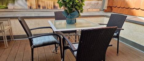 A wooden deck with wicker chairs and a glass table, overlooking a park with statues and a pond.