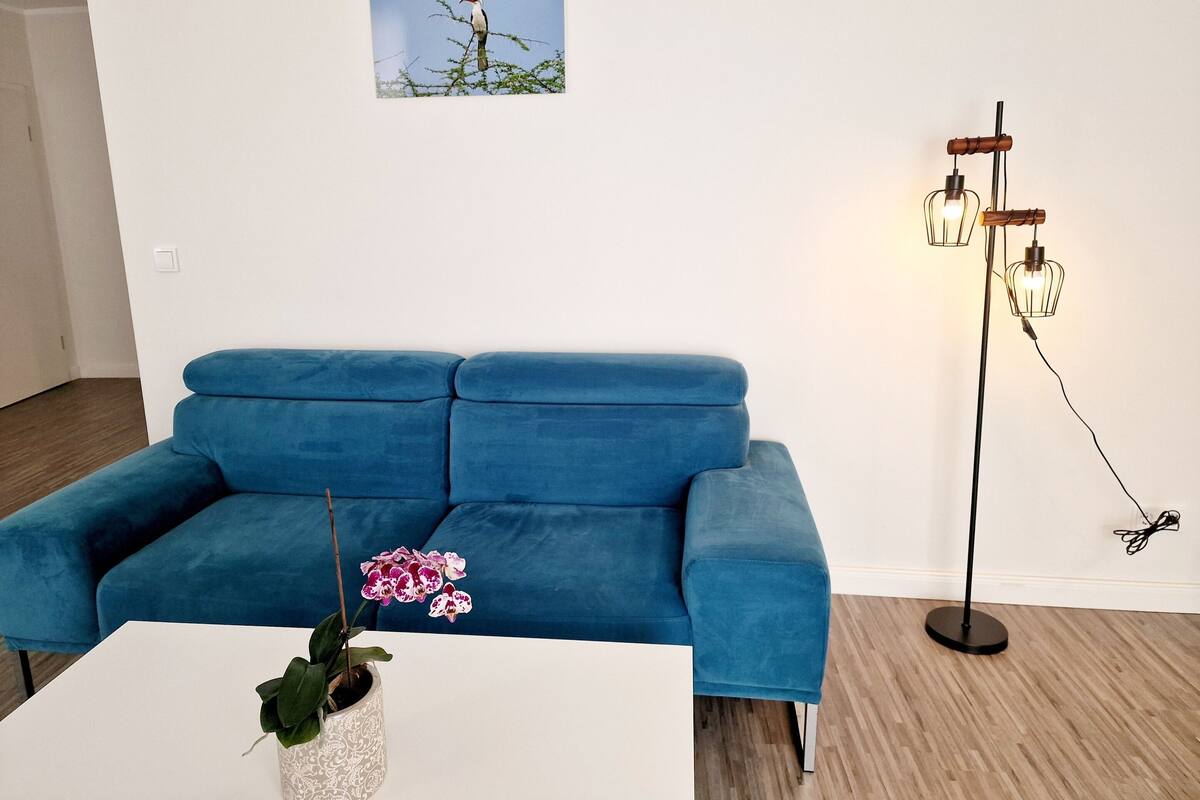 Un sofá azul, una mesa blanca con una orquídea y una lámpara de pie con tres luces.