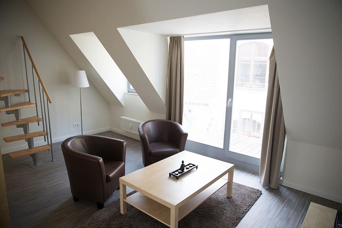 Una sala moderna con sofá, sillón y una mesa de centro. Se observa una escalera con barandilla de madera y una lámpara. La habitación tiene ventanas amplias con cortinas y vistas al exterior.