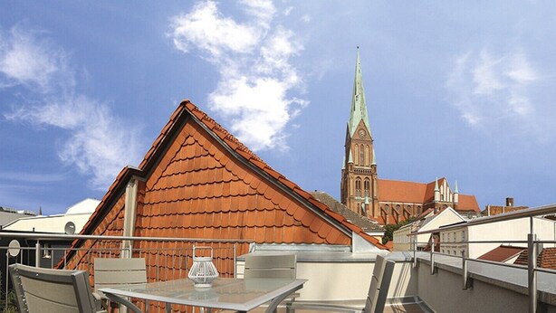 A rooftop terrace with a table and chairs, a view of a church with a tall spire, and surrounding buildings. - Domblick Schwerin (Schwerin)