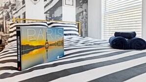 En säng med ett randigt överkast, en bok med titeln ”Paris och dess ljus” och hoprullade handdukar på en hylla bredvid.