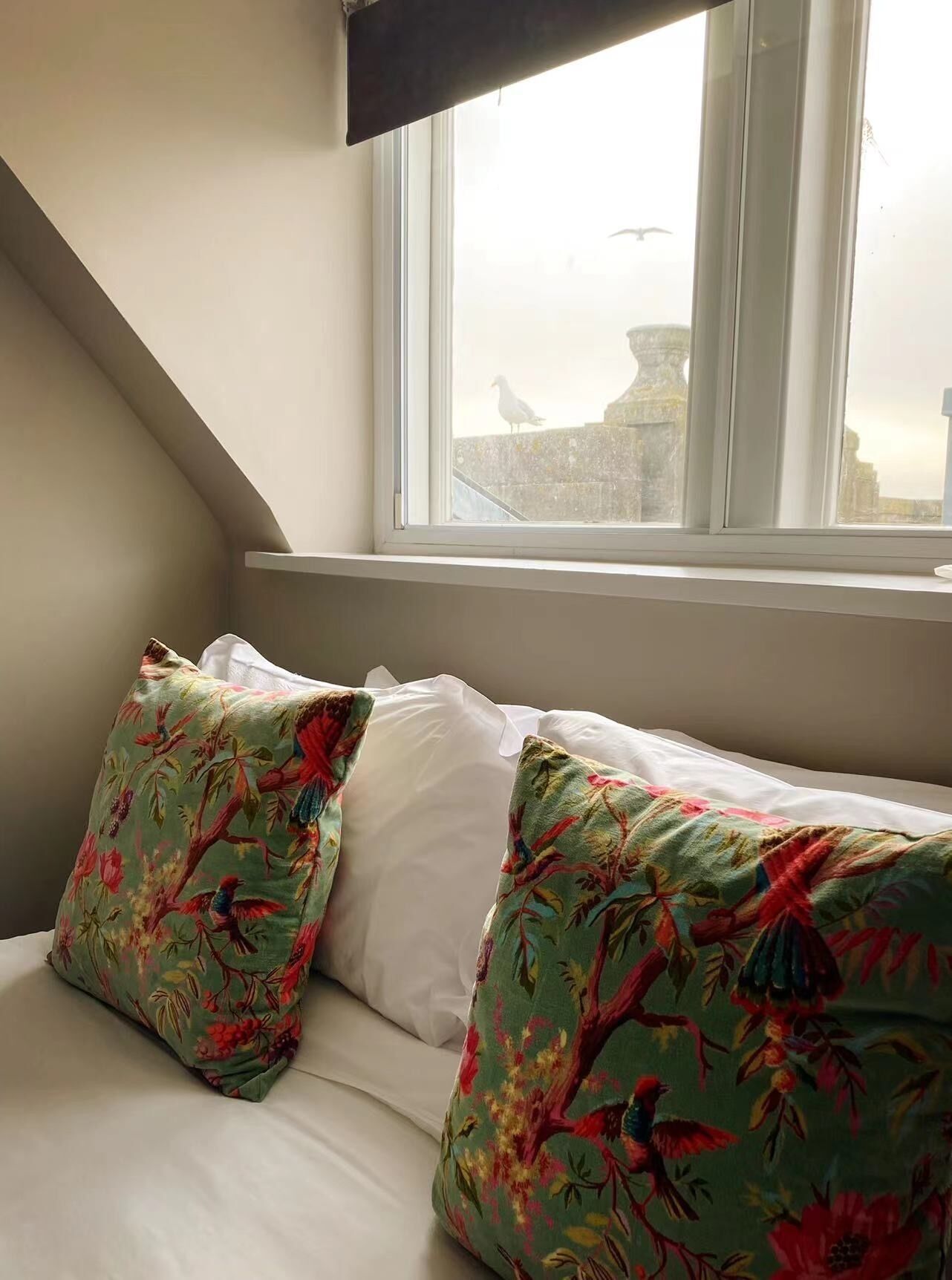 A bedroom with a sloped ceiling, a window with a view of a cityscape, and two pillows with a floral pattern.