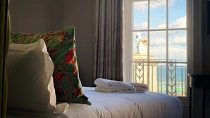 Habitación de hotel con cama, almohada estampada con flores, toallas blancas y vistas a una estatua y al océano a través de amplios ventanales.