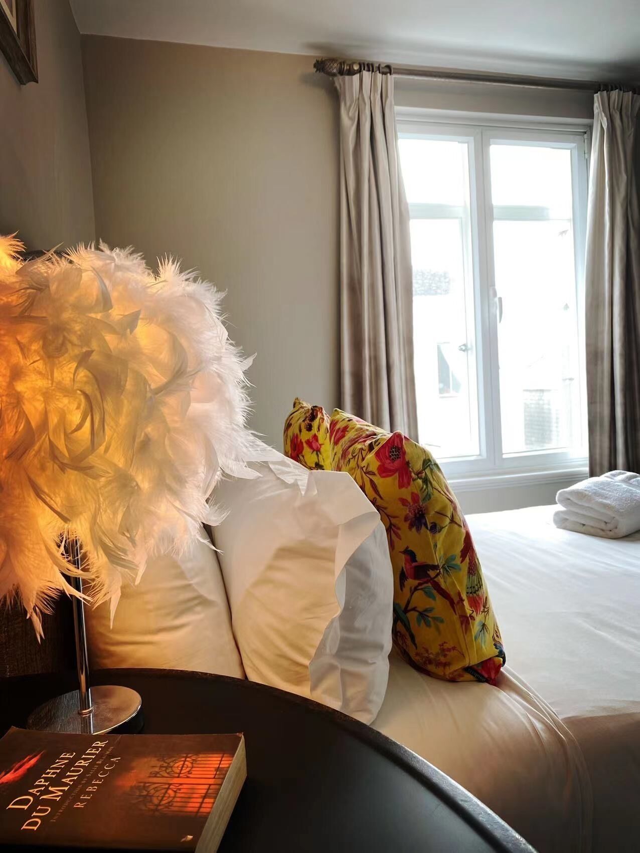 A hotel room with a bed, a lamp with a feathered lampshade, a book titled 'DAPHNE DU MAURIER', and a window with curtains.