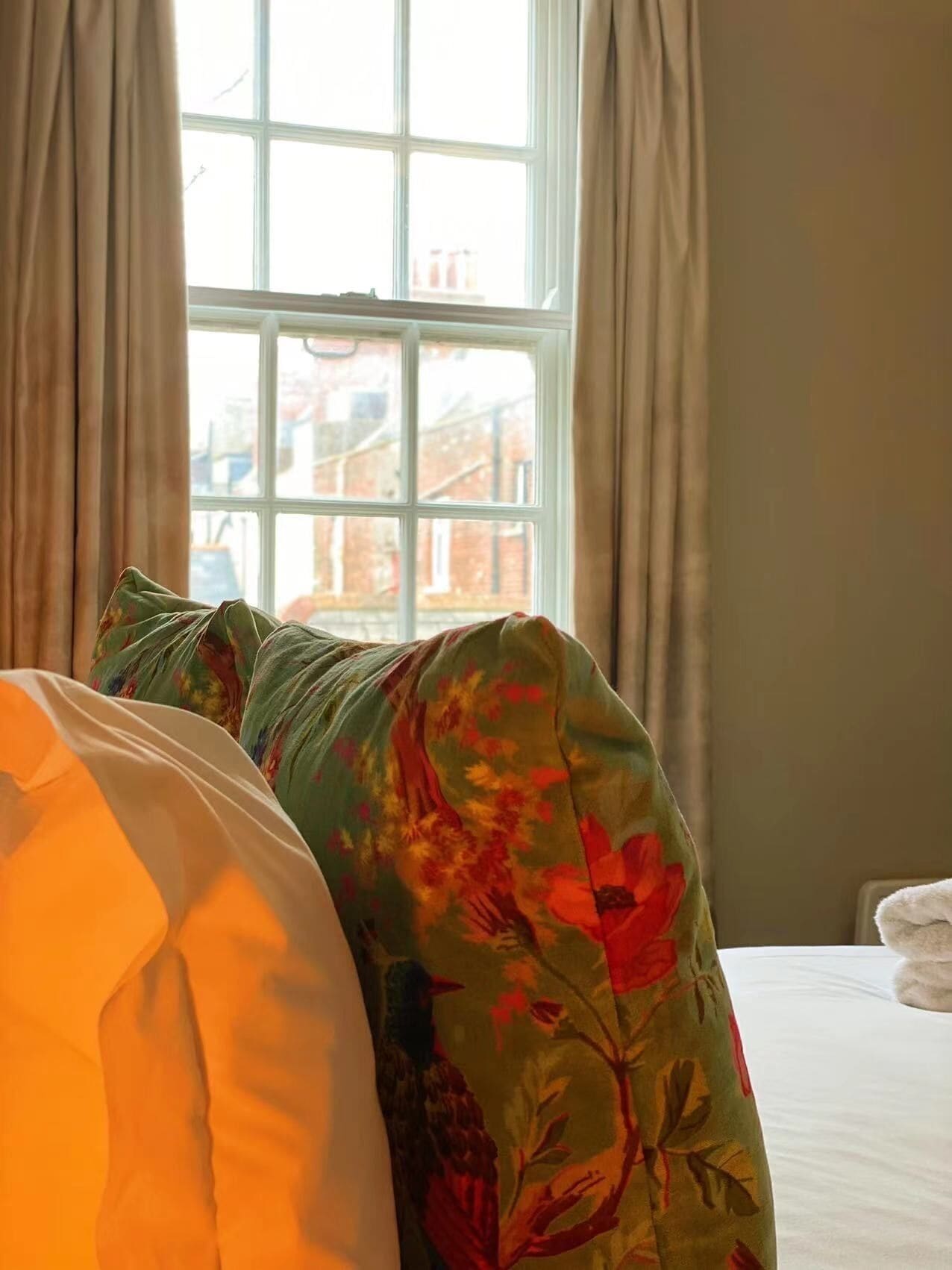 Una cama con una manta de estampado floral, una almohada blanca y vista a un edificio de ladrillo a través de una ventana con cortinas.