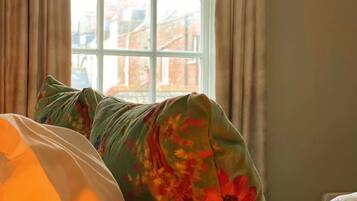 Una cama con una manta de estampado floral, una almohada blanca y vista a un edificio de ladrillo a través de una ventana con cortinas.