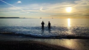 Escena de playa con dos personas de pie en aguas poco profundas, un atardecer de fondo y barcos en el horizonte.