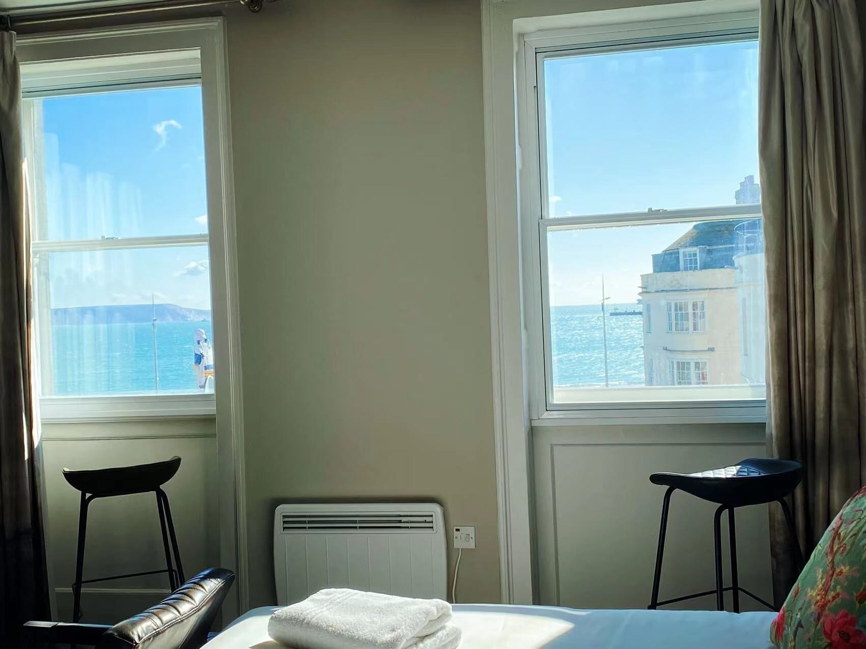 A hotel room with a bed, two chairs, a radiator, and large windows with a view of the sea and buildings.
