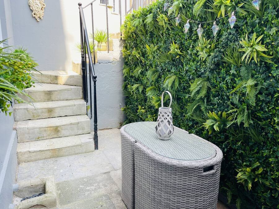 A staircase leading up to a white wall, a wicker chair, and a hedge with hanging lights.