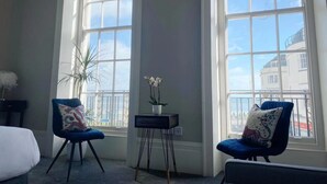 Una habitación con dos sillones azules, una mesita con un florero y grandes ventanales con vistas a edificios.