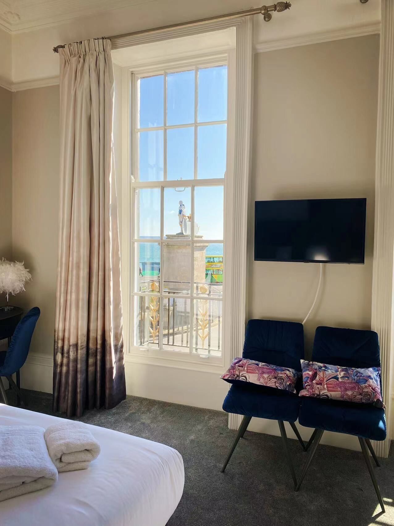 A hotel room with a bed, two blue armchairs, a TV, a window with a view of the sea, and a statue outside.