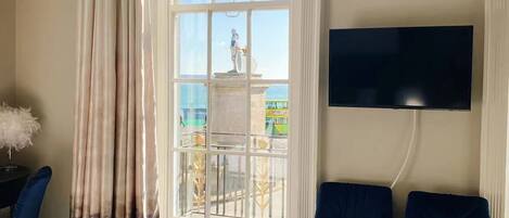 A hotel room with a bed, two blue armchairs, a TV, a window with a view of the sea, and a statue outside.
