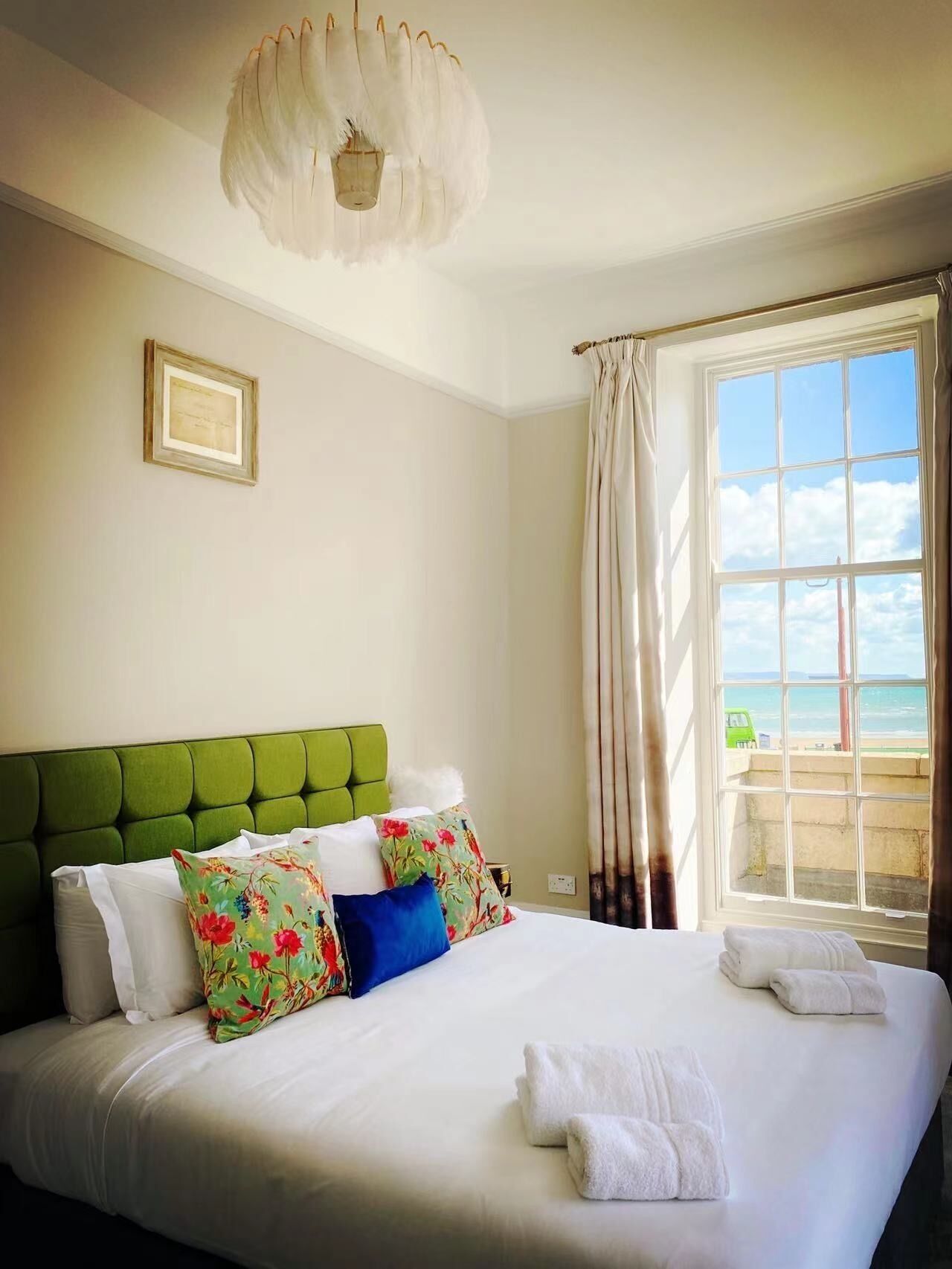 A neatly made bed with white linens and a green headboard, a framed picture on the wall, and a window with curtains.