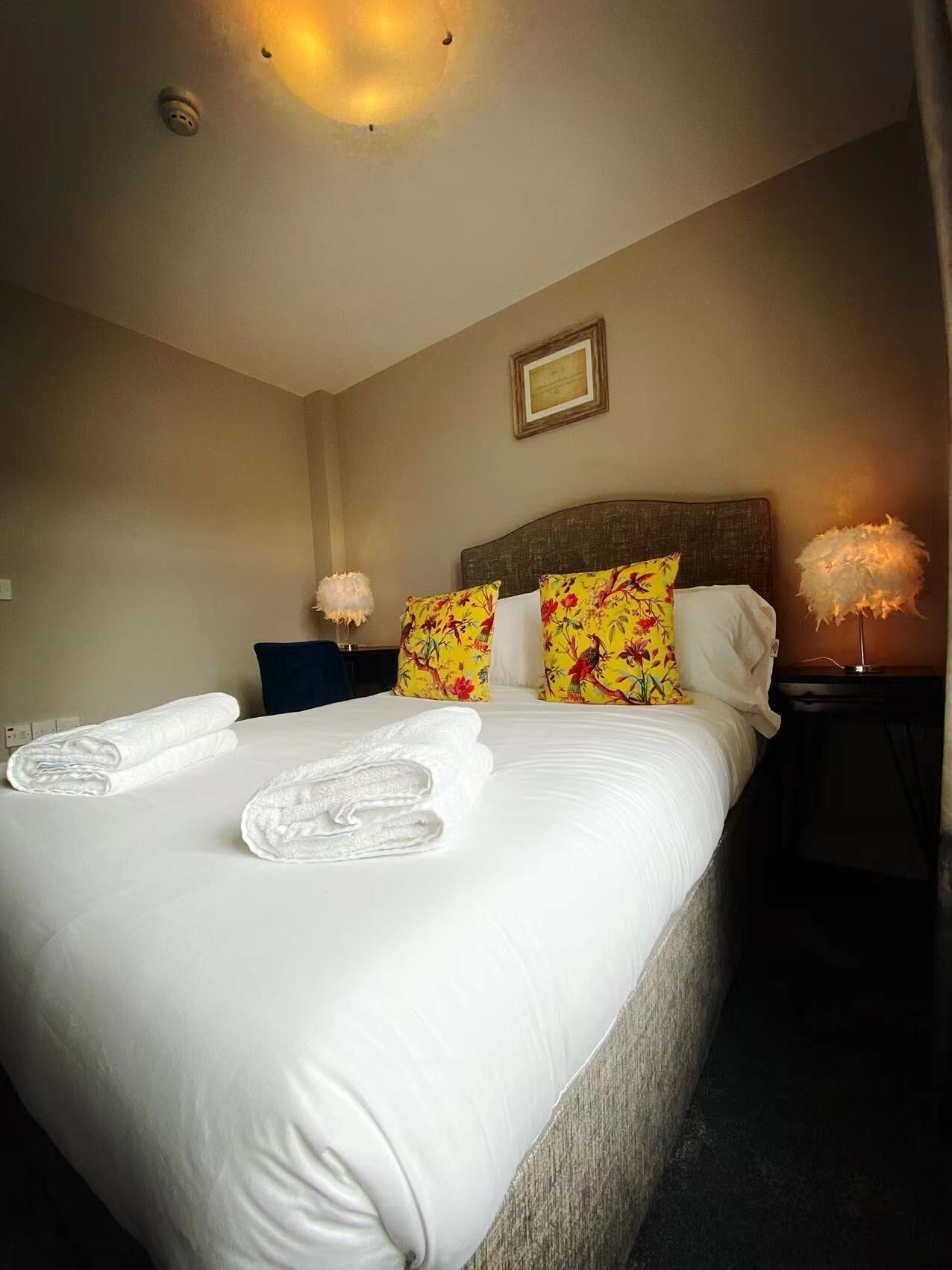 A neatly made bed with white linens and two towels, a wooden headboard, floral pillows, a bedside lamp, and a framed certificate on the wall.