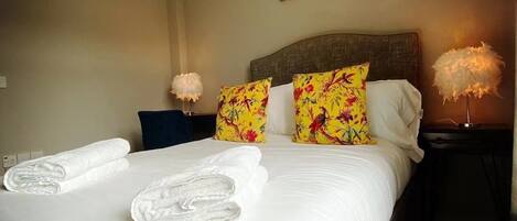 A neatly made bed with white linens and two towels, a wooden headboard, floral pillows, a bedside lamp, and a framed certificate on the wall.