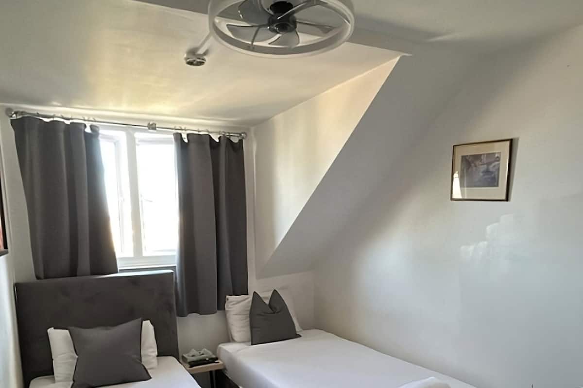 A hotel room with two beds, a ceiling fan, a window with curtains, and a small table with a fan.