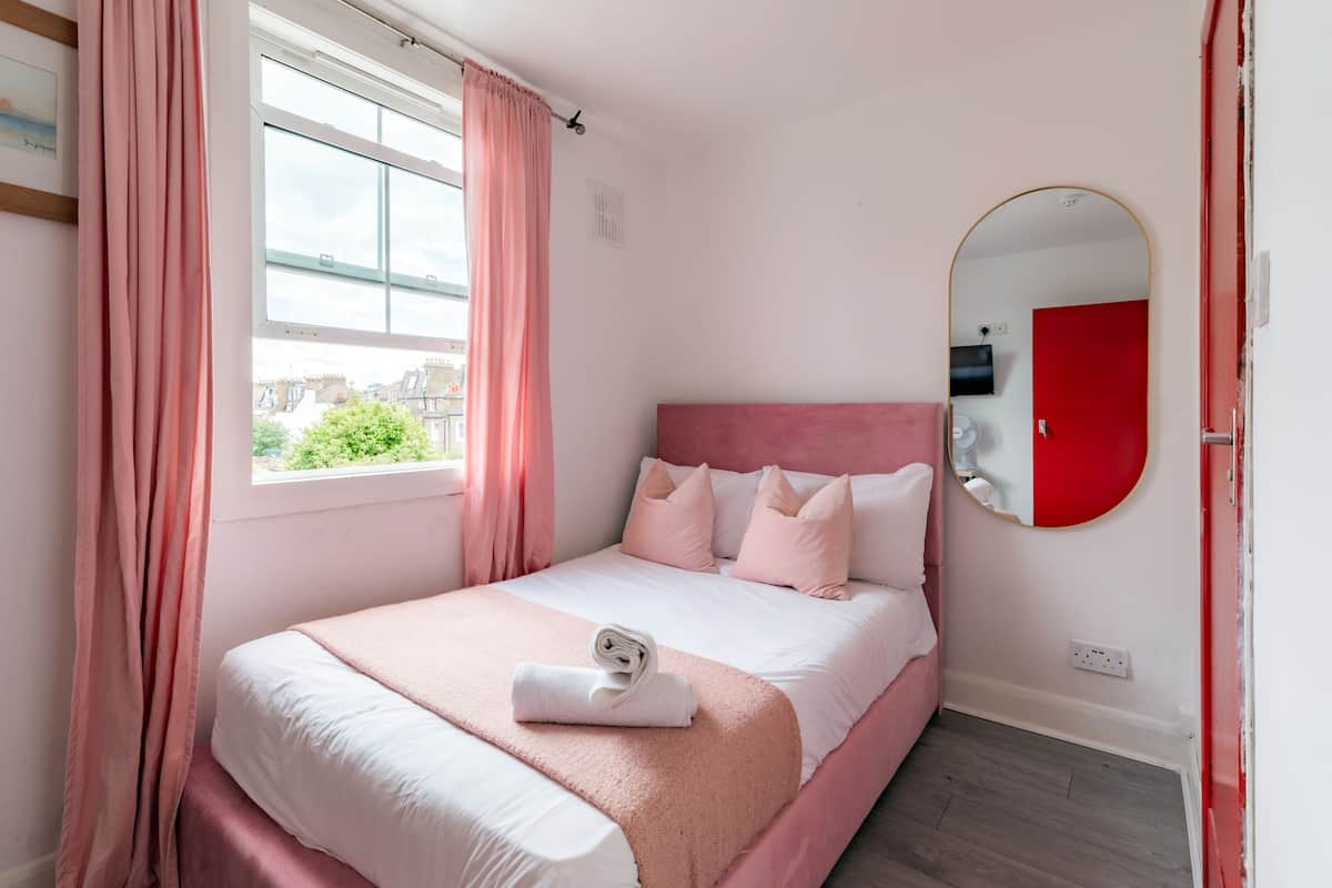 A bedroom with a bed, pink curtains, a window, a mirror, and a red door.