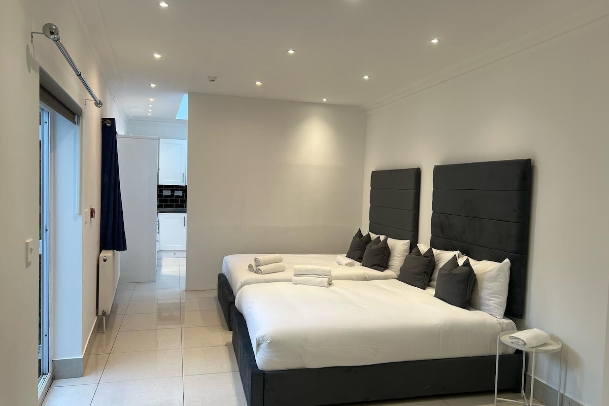 A modern bedroom with a large bed, two bedside tables, and a view of the kitchen.