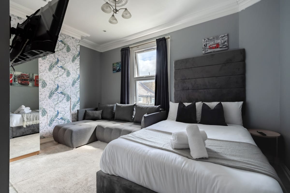A modern bedroom with a large bed, a sofa bed, and a flat-screen TV mounted on the wall.