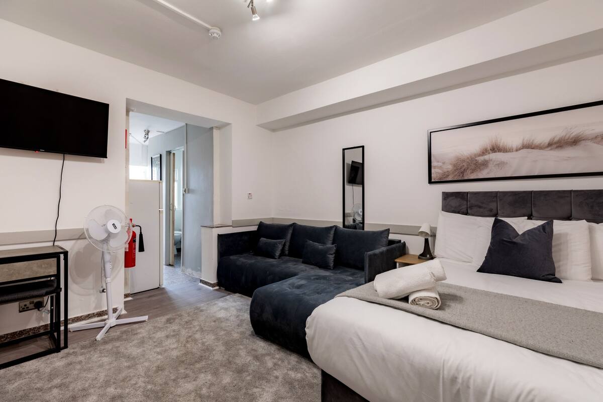 A modern hotel room with a large bed, a sofa, a flat-screen TV, a standing fan, and a painting on the wall.