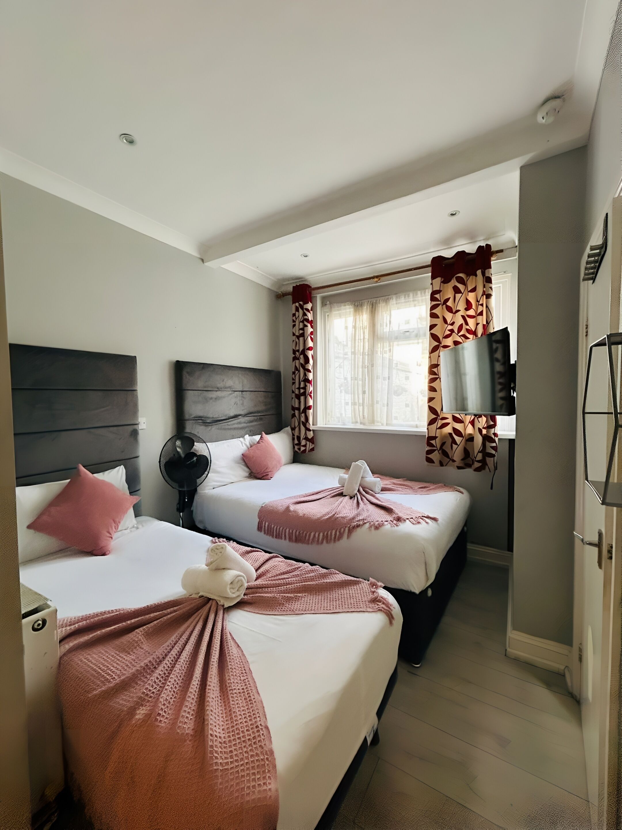 Une chambre d’hôtel avec deux lits, chacun recouvert d’une couverture et d’un oreiller roses, un ventilateur de plafond et une fenêtre ornée de rideaux à motifs.