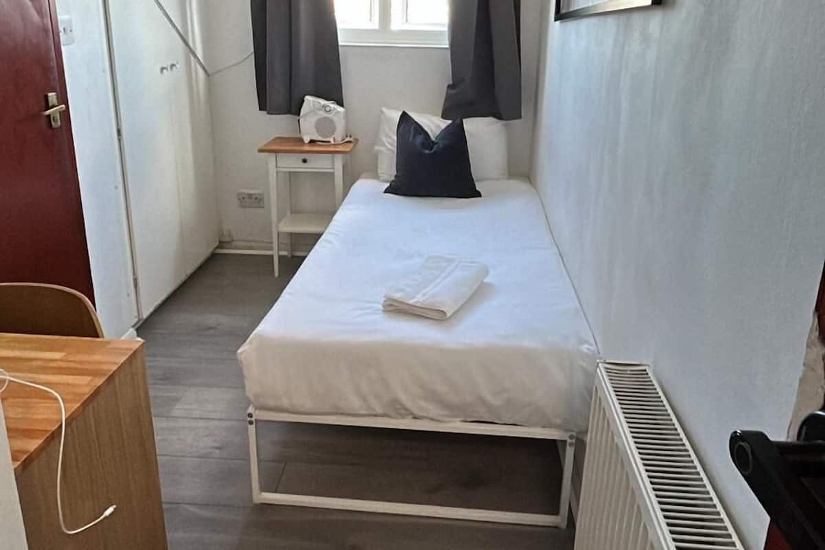A compact room with a bed, a small table, a radiator, and a ceiling fan.