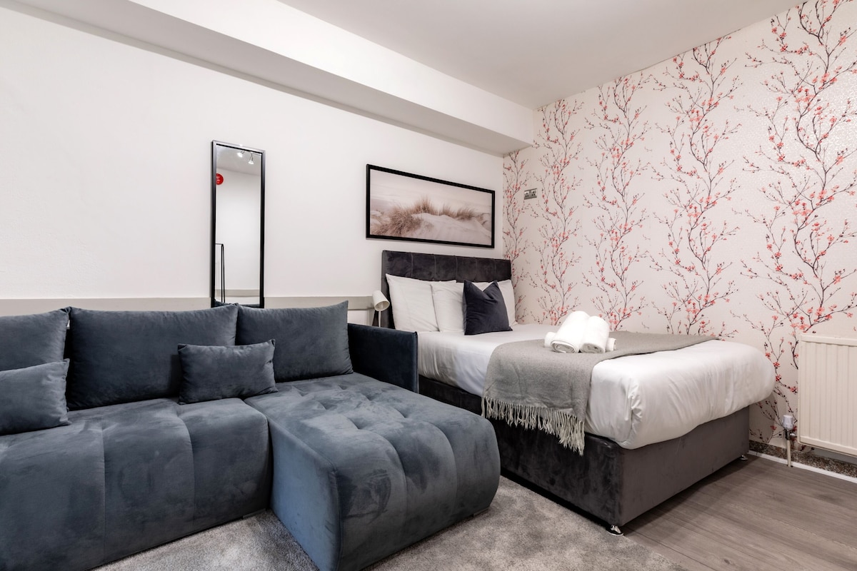 A modern bedroom with a grey sectional sofa, a single bed with a grey duvet, a bedside table with a lamp, a framed picture on the wall, and a mirror.