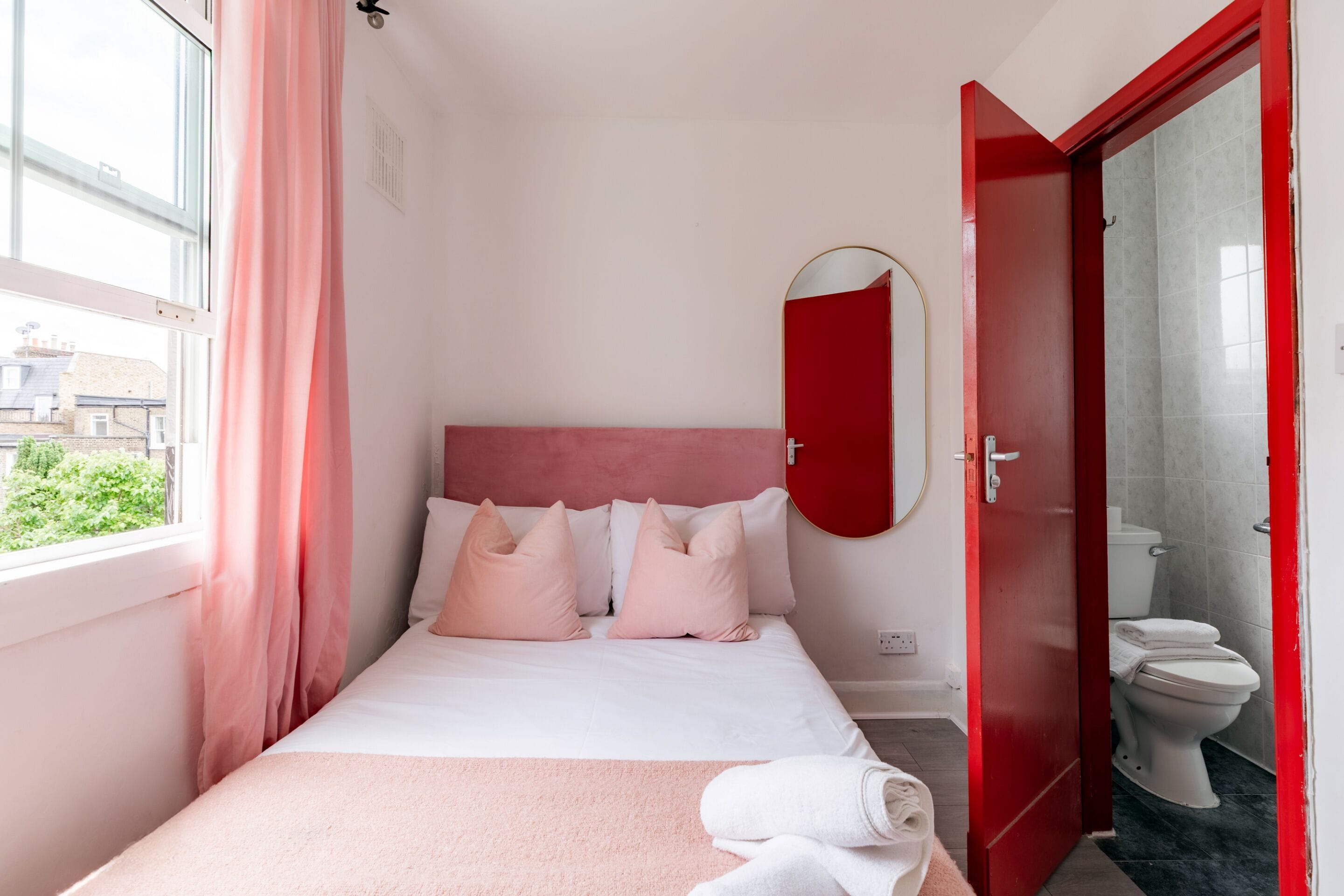 Habitació d'hotel amb un llit, una finestra amb cortines, una porta vermella i un vàter visible a través de la porta.