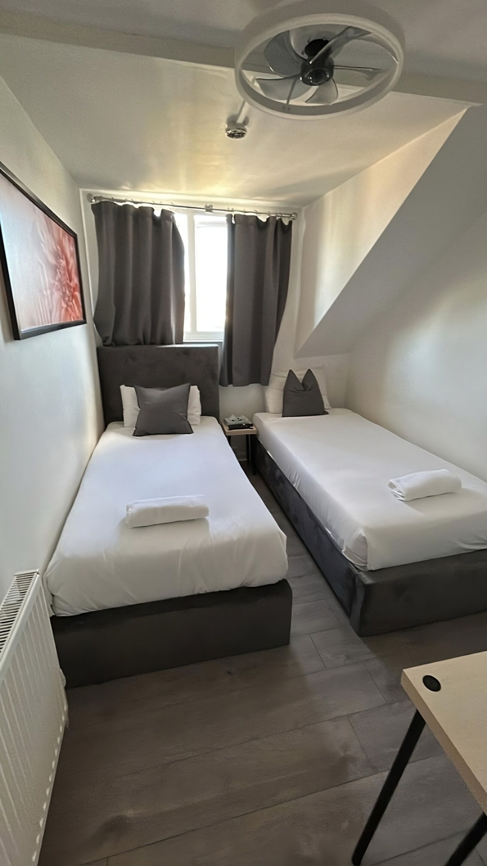 A compact hotel room with two single beds, a small table, a ceiling fan, and a window with curtains.