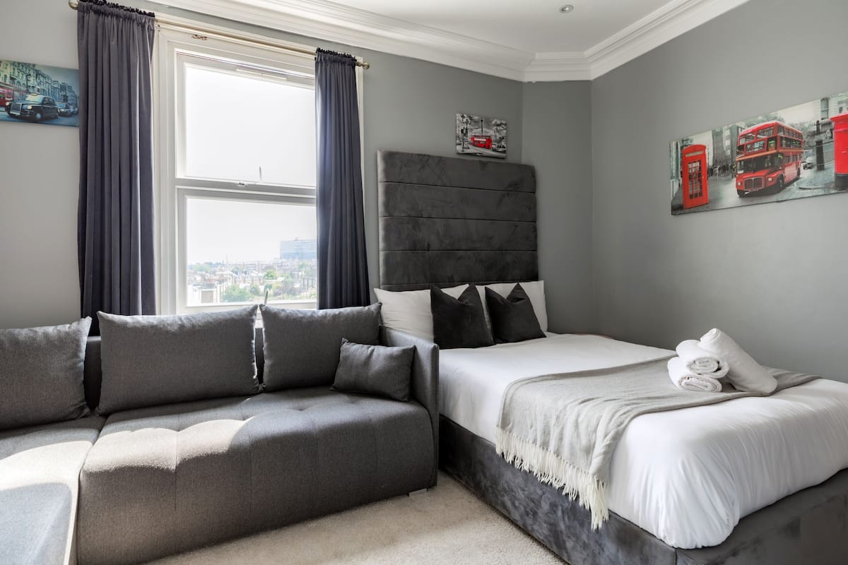 A modern bedroom with a grey sofa, a bed with white linens, and a cityscape view through the window.