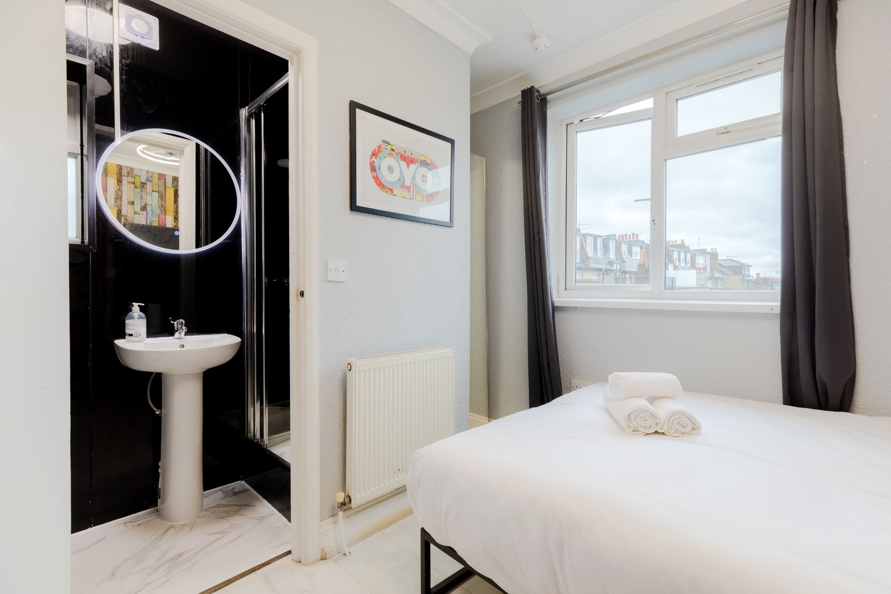 Habitación de hotel con cama, lavamanos blanco, espejo, cuadro enmarcado y ventana con cortinas.
