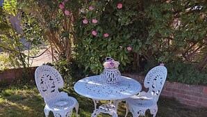 A white outdoor seating area with a round table and two chairs, surrounded by greenery and pink flowers. - The Cheese House (Salisbury)