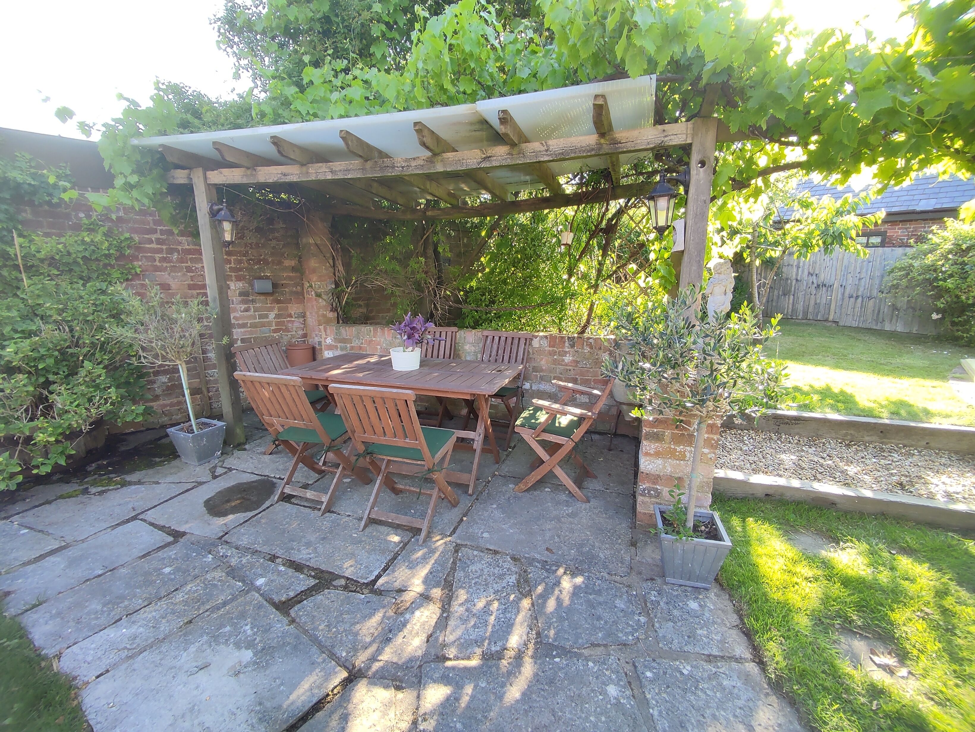 A wooden pergola with a table and chairs underneath, surrounded by greenery and a brick wall.