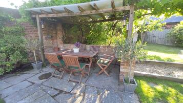 A wooden pergola with a table and chairs underneath, surrounded by greenery and a brick wall.
