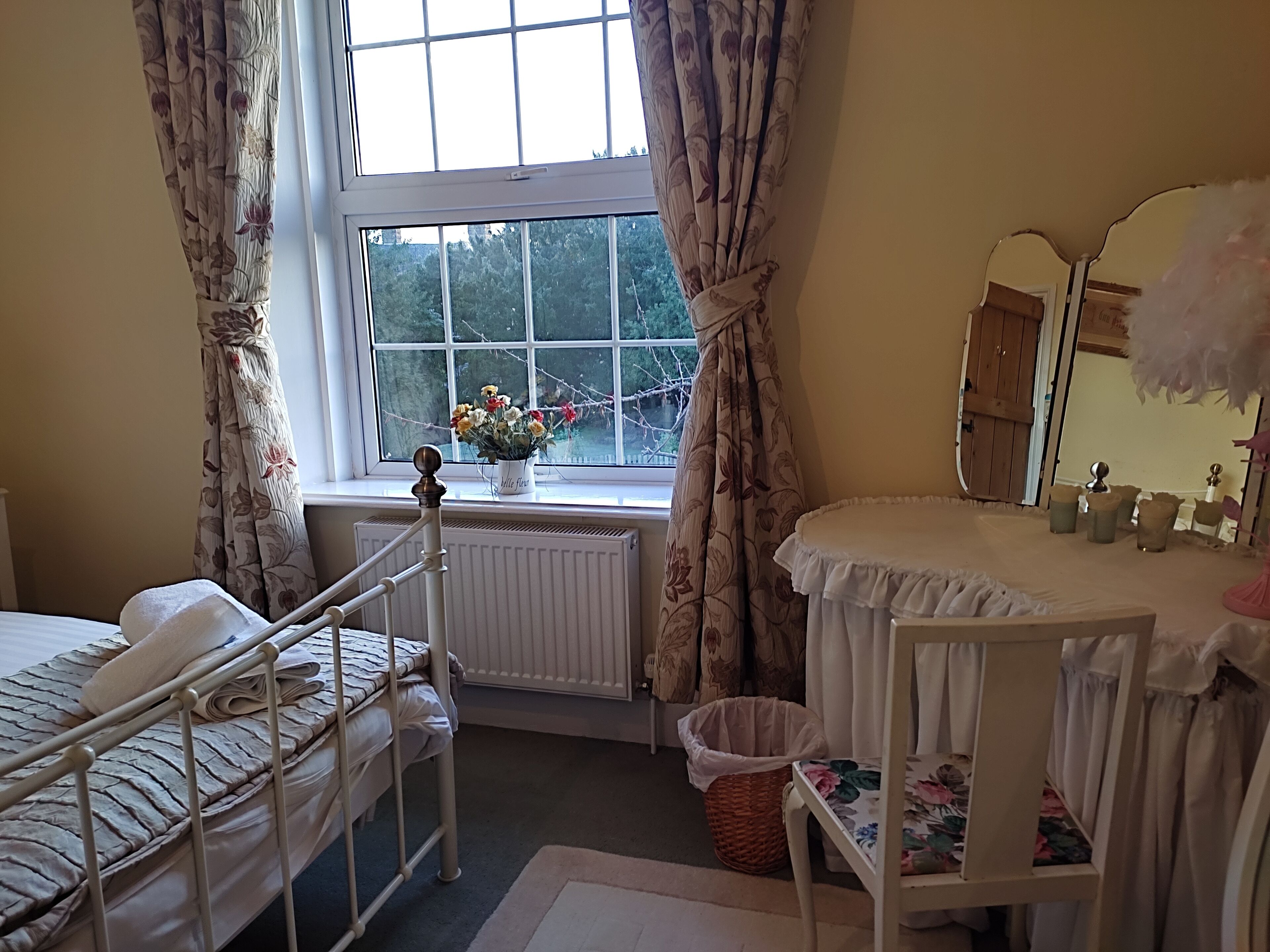 A bedroom with a bed, a window with curtains, a radiator, a mirror, and a bedside table.