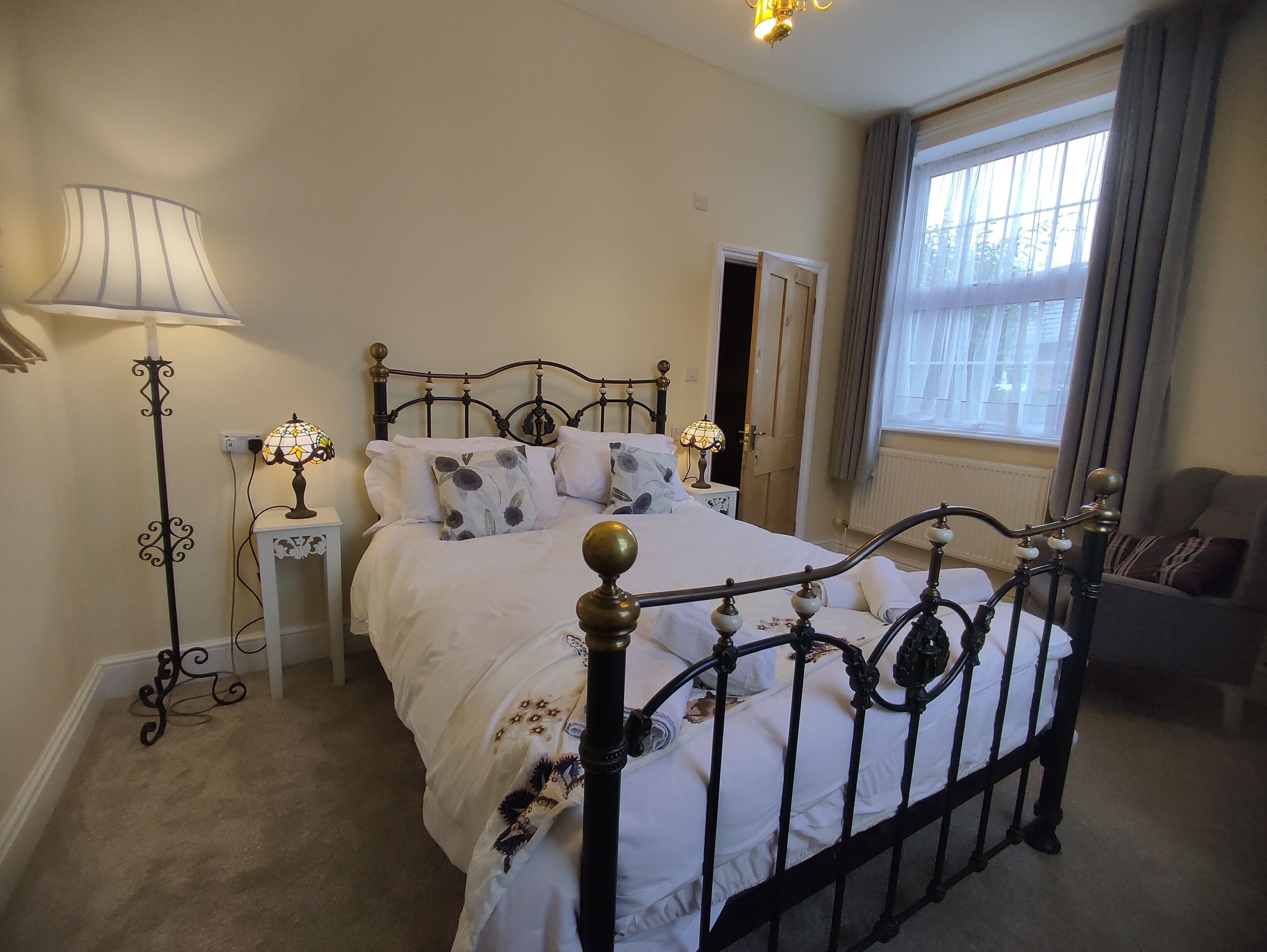 Een slaapkamer met een hemelbed, nachtkastje met lamp, een dressoir en een raam met gordijnen.
