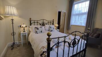 Een slaapkamer met een hemelbed, nachtkastje met lamp, een dressoir en een raam met gordijnen.