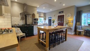 A kitchen with a central island, a stove, and a microwave. There is a clock on the wall and a door leading to another room. - The Cheese House (Salisbury)