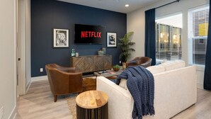 A modern living room with a sofa, two armchairs, a coffee table, and a TV mounted on the wall displaying 'NETFLIX'.