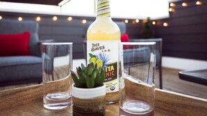 A bottle of Tres Agaves Itaniactic on a wooden table with two glasses and a small potted plant.