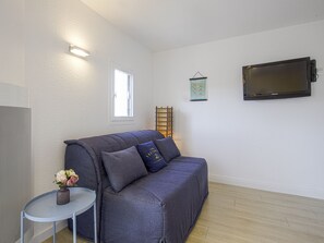 A modern living room with a grey sofa, a small side table with a vase of flowers, a mounted flat-screen TV, and a window with blinds. - Studio des Falaises - Welkeys (Anglet)