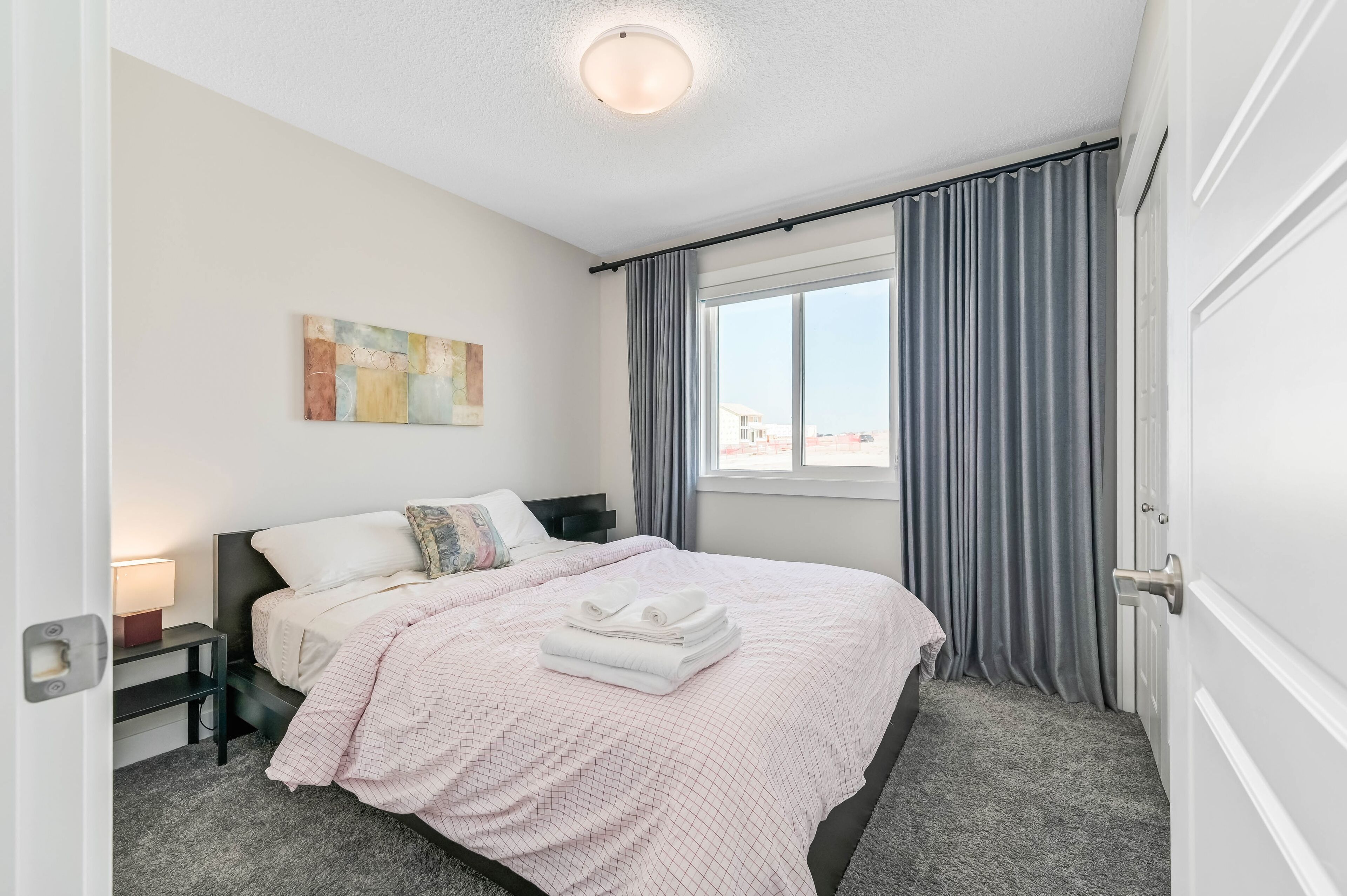 A neatly made bed with a pink checkered bedspread, a folded white towel, and a nightstand with a lamp.