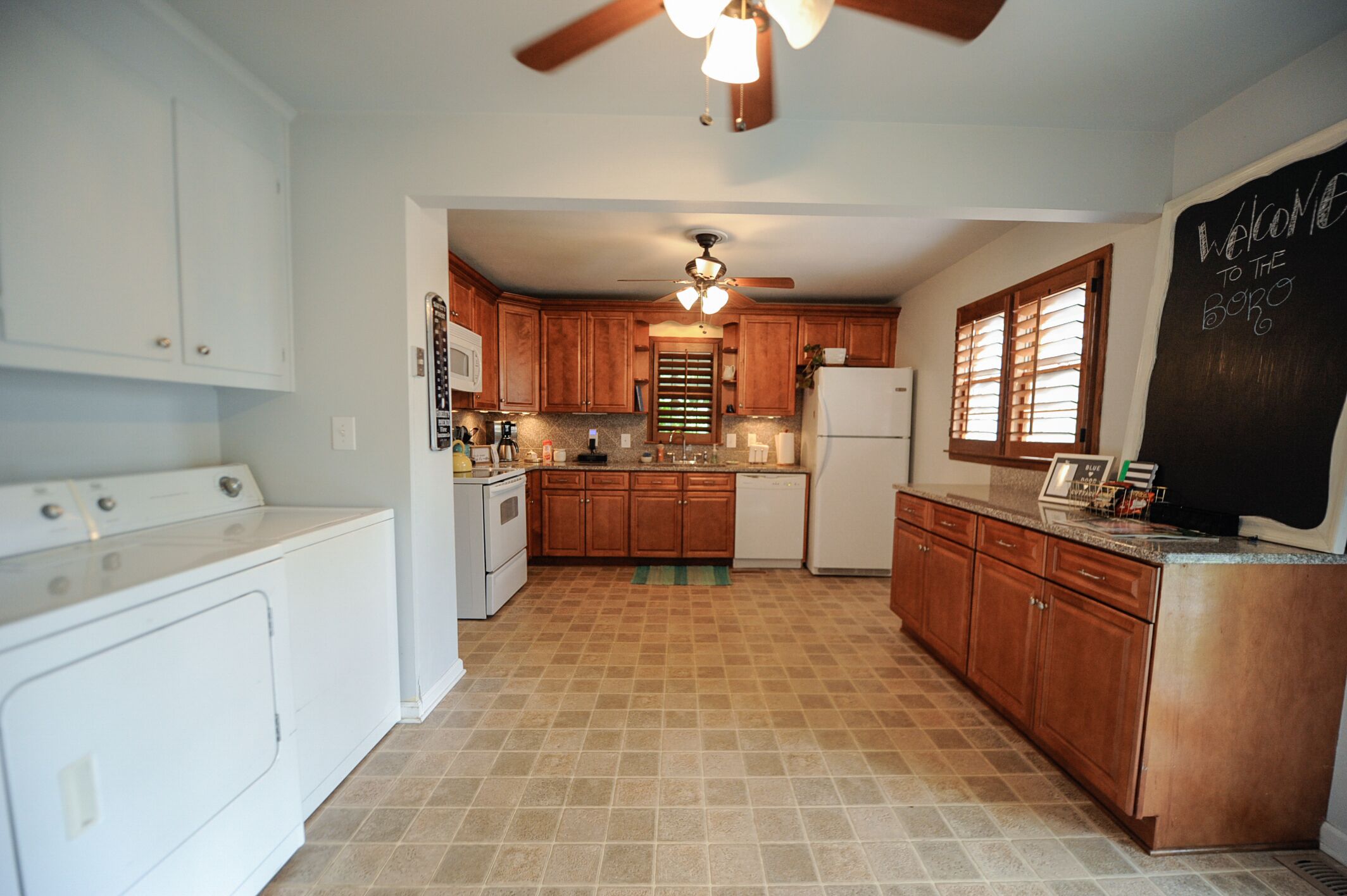 A kitchen with wooden cabinets, a ceiling fan, a refrigerator, and a chalkboard with a welcome message.