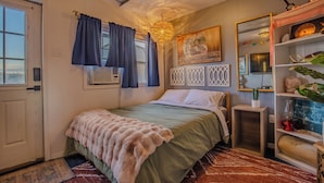 Un dormitorio con una cama, una mesita de noche, una estantería decorada y una ventana con cortinas.
