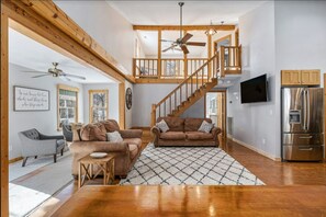 A spacious living area with a wooden staircase, a ceiling fan, and a modern kitchen with a stainless steel refrigerator.