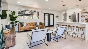 A modern kitchen with a central island, bar stools, and a sofa in a bright living area with large windows.