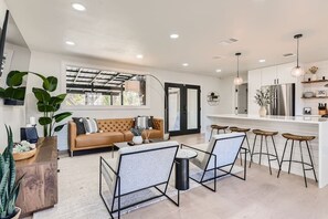 A modern kitchen with a central island, bar stools, and a sofa in a bright living area with large windows.