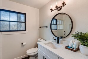 A bathroom with a toilet, a sink, a mirror, a window, and a wall-mounted light fixture.