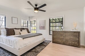 A bedroom with a bed, a wooden dresser, a ceiling fan, and large windows.